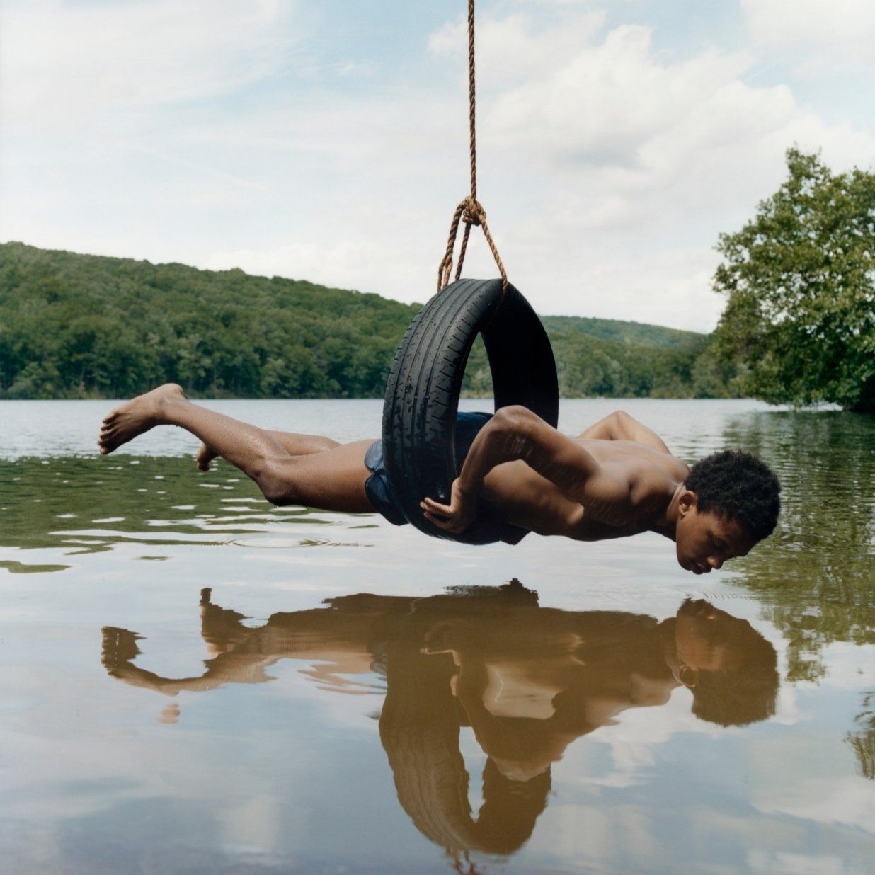 A young Black man wearing swim trunks is suspended horizontally in a tire swing over a muddy river, with trees in the background.