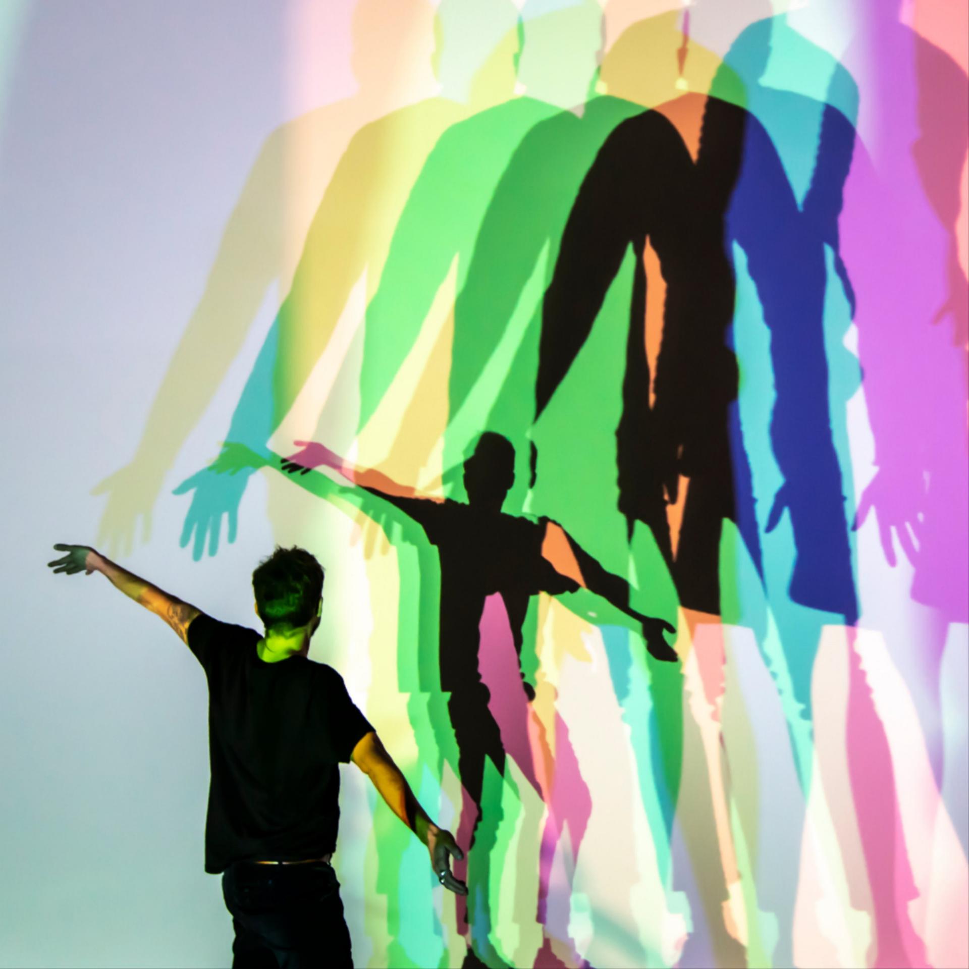 A person and their shadow repeated in many colors stands in front of a wall where other shadows in many colors are projected
