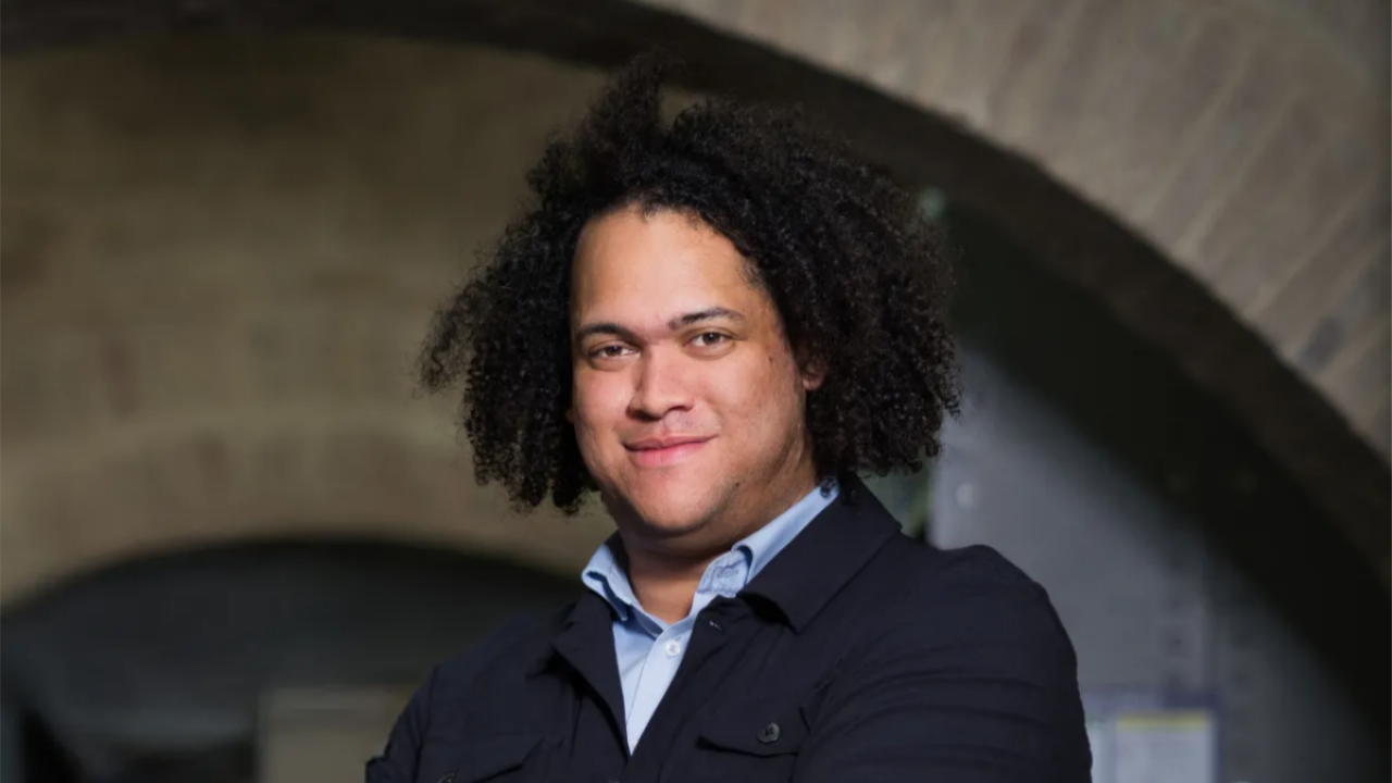 A man with medium-length dark curly hair, wearing a pale blue button-won shirt and a dark jacket, smiles slightly with his arms crossed beneath a stone archway.